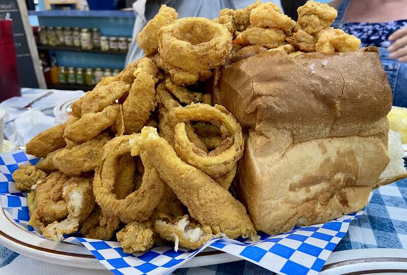 A fried seafood platter with onion rings and a loaf of French bread on blue-checkered paper.